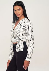 Camicia avvolgente bianca con motivo a strisce nere, dotata di maniche lunghe e dettagli in vita annodati, abbinata a pantaloni neri aderenti.