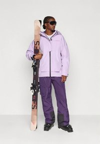 Lilla skijakke med hætte, sorte accenter og matchende lilla skibukser. Holder ski med bindinger. Solbriller fuldender outfittet.