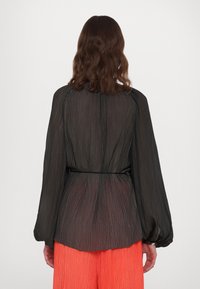 Blouse noire à manches longues plissée avec une ceinture à nouer à la taille, associée à un pantalon texturé orange vif. Tissu lisse et léger.
