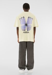 Yellow oversized t-shirt features a large purple butterfly graphic on the back, with black text below. Paired with gray baggy pants and white shoes.
