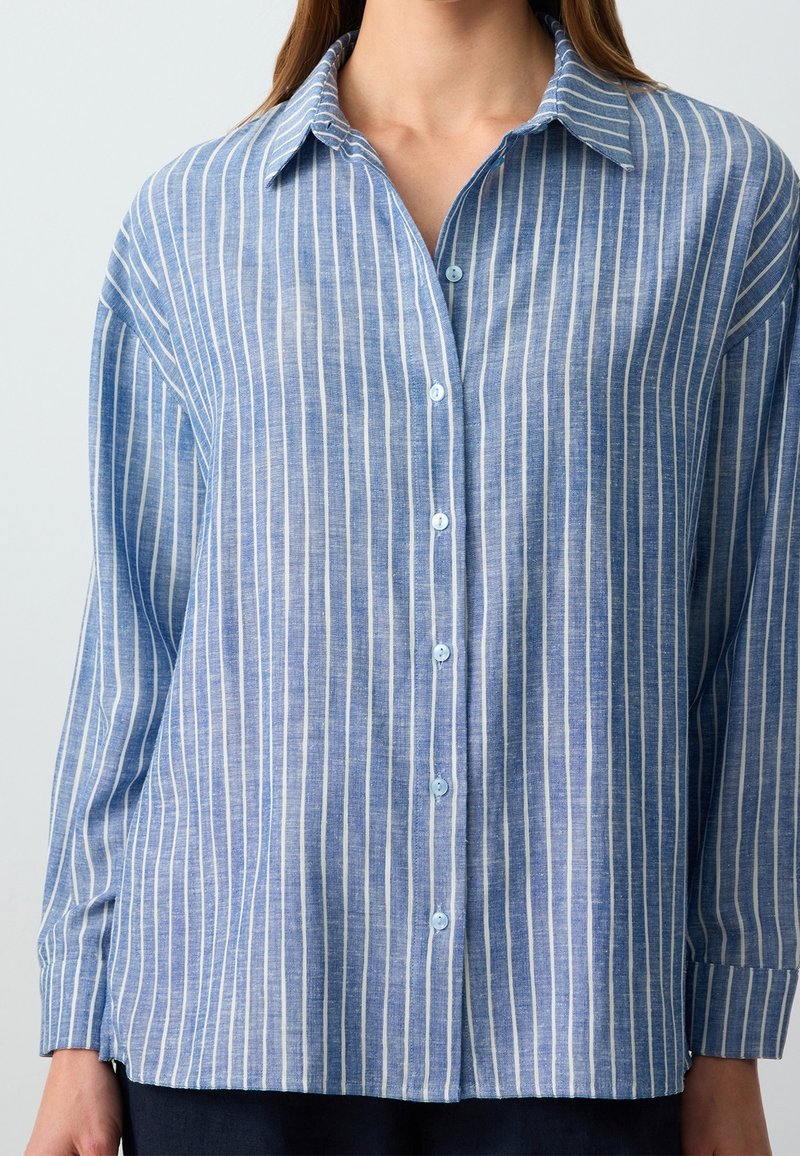 Chemise bleue boutonnée avec des rayures blanches verticales, manches longues et col classique portée par une personne.