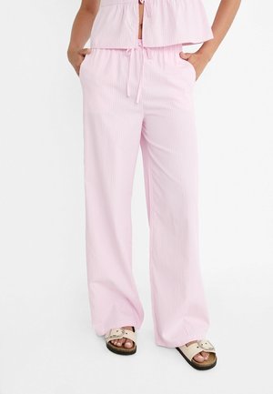 Persoon draagt lichtroze gestreepte broek met wijde pijpen en een bijpassend topje, met de handen in de zakken, gecombineerd met beige sandalen met gesp.