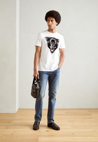 Camiseta blanca con un gráfico de pantera negra, combinada con vaqueros azules y mocasines negros, sosteniendo un pequeño bolso negro.
