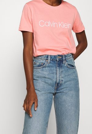 Persoon draagt een roze Calvin Klein T-shirt, ingestopt in lichtblauwe high waist spijkerbroek, met een hand ontspannen langs het lichaam.