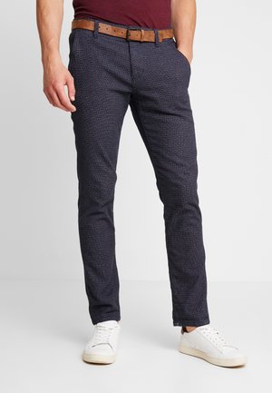 Mann trägt dunkelblaue, strukturierte Slim-Fit-Hose mit braunem Ledergürtel, weißen Sneakers und kastanienbraunem Hemd, steht mit einer Hand in der Hosentasche.