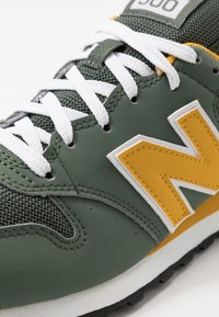 Gröna lädersneakers med meshdetaljer, vita snören, gult "N"-logga och svart gummisula. Har förstärkta sömmar och vadderad krage.