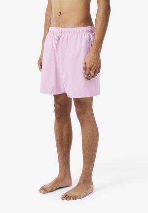 Mann trägt hellrosa Schnürzug-Shorts und steht barfuß vor einem schlichten weißen Hintergrund.