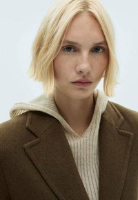 Hnedý vlnený kabát so štruktúrovaným klopom, preložený cez svetlo béžovú pletenú mikinu s kapucňou. Model má krátke blond vlasy a jasné modré oči.