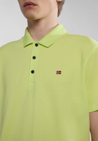 Limetkově zelená polo tričko vyrobené z texturované látky, s límečkem, třemi černými knoflíky a malým vyšitým detailem vlajky na hrudi.