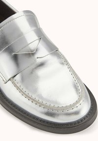 Mocassin en cuir argenté avec un bout arrondi, panneau décoratif cousu, semelle plate et texture lisse. Couture renforcée le long des bords.