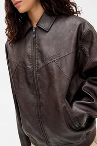 Žena v tmavě hnědé kožené zipové bundě s vyraženým nápisem "BDG JEANS" na hrudi, s rukama v kapsách a volnými vlnitými vlasy.