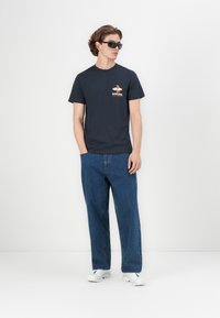 Mørkeblå bomull t-skjorte med grafisk design på fronten, sammen med løstsittende denimjeans. Modell iført mørke solbriller og hvite joggesko.
