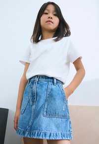 Falda de mezclilla con dos bolsillos delanteros, lavado azul claro, dobladillo deshilachado, combinada con una camiseta blanca lisa. Diseño simple y casual.