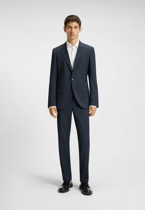 Navyblauer Anzug mit tailliertem Schnitt, mit einem einzigen Knopfverschluss, Reverskragen und flachen Hosen. Weißes Hemd und schwarze Schuhe.