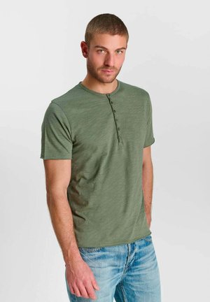 Mann trägt ein olivgrünes Henley-Shirt mit kurzen Ärmeln und hellblaue Jeans und steht vor einem schlichten hellen Hintergrund.