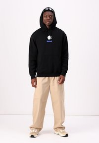 Black hoodie with a front pocket and blue "Kodiak" text. Paired with beige pants and light sneakers. Minimalist design, relaxed fit.