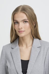 Jonge vrouw met lang blond haar, gekleed in een lichtgrijze blazer over een zwart shirt, die met een neutrale expressie recht in de camera kijkt.