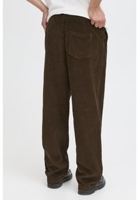 Brown corduroy pants with a relaxed fit, visible vertical ribbing, and a back pocket. Worn with a plain white top and black shoes.
