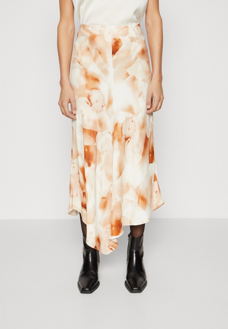 Femme portant une jupe midi à motif abstrait blanc et orange rouille, associée à des bottines noires, se tenant devant un fond uni.