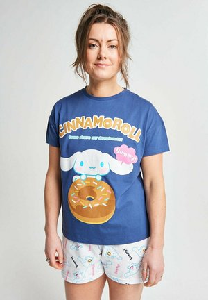 Frau trägt ein blaues Cinnamoroll-T-Shirt mit Donut-Grafik und passende weiße Shorts mit Hund- und "yummy"-Drucken, steht vor einfachem Hintergrund.