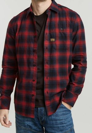 Mężczyzna ubrany w czerwoną i czarną kratowaną koszulę z guzikami na czarną koszulkę i niebieskie dżinsy, z ręką w kieszeni, na jednolitym tle.