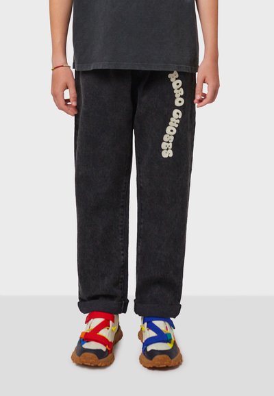 Pantalones de mezclilla negros con un corte relajado y texto vertical en blanco "RUG CHOSES" en la pierna izquierda. Un par de zapatillas multicolores con suelas gruesas.