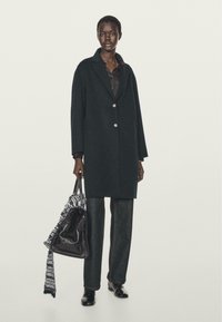 Lang donkere jas met een kraag, sluiting met één knoop, gedragen over een zwart shirt. Model houdt een zwarte tas vast met een geruite sjaal die eroverheen hangt.