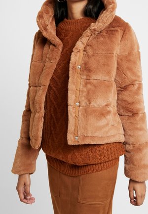 Kvinde iført en lysebrun blød hættejakke over en rustfarvet kabelstrikket sweater og en brun ruskindsnederdel.