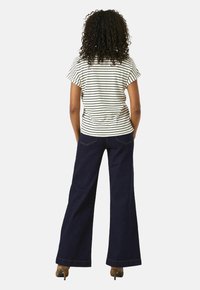 Persoon met krullend haar, gekleed in een wit en zwart gestreept shirt met korte mouwen, donkere wijde jeans en bruine hakken, staande met de rug naar ons toegekeerd.