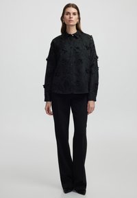 Zwarte shirt met bloemen uitsparingen, knoopsluiting, lange mouwen, gestructureerde stof, gecombineerd met op maat gemaakte zwarte broek en puntige schoenen.