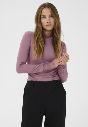 Kvinde med langt hår iført en mauve ribbet langærmet top med blonder ved ærmerne og sorte højtaljede bukser foran en ensfarvet hvid baggrund.