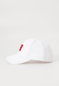 White cotton baseball cap with a red rectangular patch featuring black lettering. The cap has a curved brim and embroidered eyelets on the top.