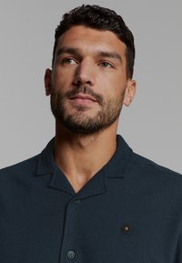 Man met kort, krullend haar en een baard, gekleed in een donkerblauw overhemd met kraag en een zwart hexagonaal logo op de borst, kijkt iets omhoog en naar de zijkant.