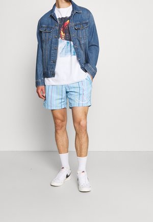 Veste en denim, t-shirt graphique blanc, shorts de bain rayés bleu clair, chaussettes blanches et baskets blanches avec des accents noirs.