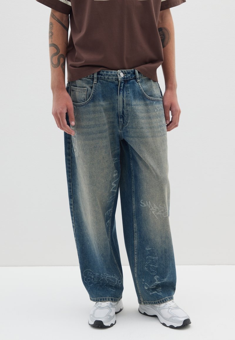 Jeans de corte holgado, de color azul desvanecido, fabricados en tela de vaquero, con ligeras desgastaciones y gráficos similares a graffiti en las piernas inferiores. Combinados con zapatillas blancas.