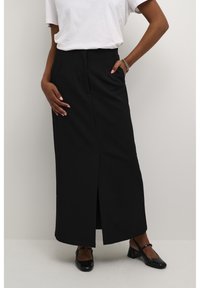 Jupe maxi noire avec une fente à l'avant, en tissu lisse. Équipée de passants de ceinture et d'une taille ajustée, associée à des chaussures noires.
