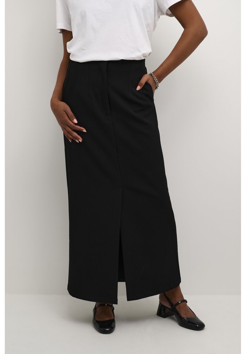 Jupe maxi noire avec une fente à l'avant, en tissu lisse. Équipée de passants de ceinture et d'une taille ajustée, associée à des chaussures noires.