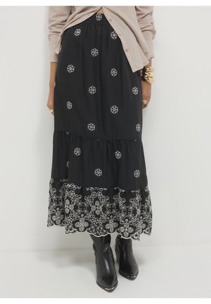 Jupe midi noire à volants avec broderie florale blanche, portée avec des bottes noires pointues et un cardigan beige à boutons.