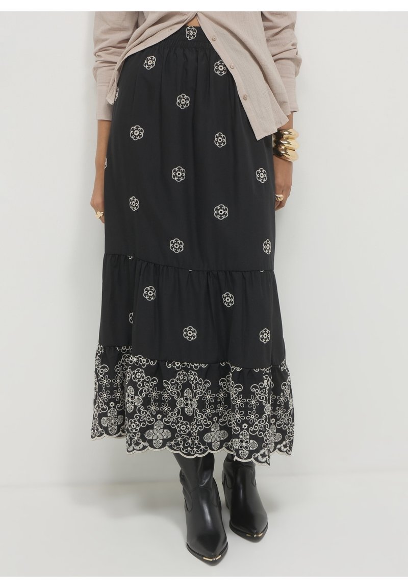 Jupe midi noire à volants avec broderie florale blanche, portée avec des bottes noires pointues et un cardigan beige à boutons.