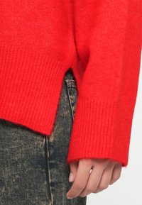 Rode wollen trui met ribgebreide manchetten, voorzien van een zijsplit. Gekleed over donker gestructureerde denim broek, wat de kleurcontrast benadrukt.