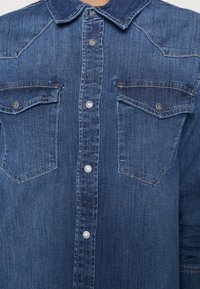 Mörkblå denimskjorta med knappstängning fram, spetsig krage och två bröstfickor. Den har silverknappar och kontrasterande sömmar.