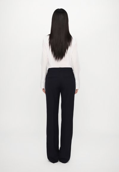 Witte blouse met lange mouwen en marineblauwe wijde broek, gladde stof, op maat gemaakt met achterzakken, model gezien van achteren tegen een eenvoudige achtergrond.