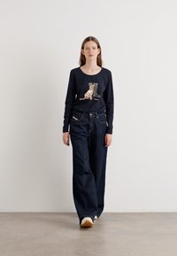 Langärmliges navy Hemd mit Grafiken, kombiniert mit weiten, dunklen Jeans und hellen Schuhen, stehend auf einem neutralen Boden.