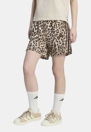 Persona che indossa pantaloncini beige con stampa leopardata, calzini bianchi con logo nero, scarpe da ginnastica beige e top beige, in piedi davanti a uno sfondo semplice.