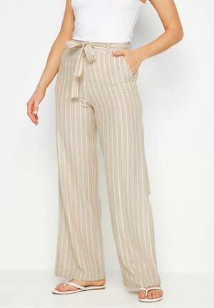 Pantalon beige à jambes larges avec des rayures verticales blanches et une ceinture à nouer, porté avec des sandales blanches type tongs et un haut blanc sans manches.