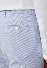 Pantaloni a righe blu chiaro e bianche con una tasca posteriore con chiusura a bottone, tessuto morbido e vestibilità sartoriale. Accompagnati da una camicia bianca.
