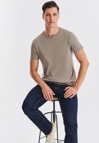 Beżowa koszulka z krótkim rękawem, okrągły dekolt, w połączeniu z ciemnoniebieskimi dżinsami. Model siedzi na stołku, ma na sobie jasne trampki.