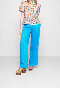 Bloes met bloemenpatroon en pofmouwen, gecombineerd met heldere blauwe wijde broek en bruine houten klompen met hak.