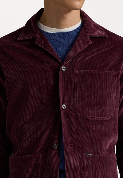 Burgundy corduroy jacket with notched collar, front pockets, and button closure. Worn over a navy knit sweater with crew neck.