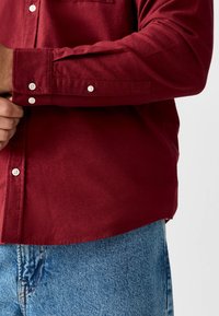 Chemise bordeaux boutonnée avec un tissu texturé, des boutons blancs et une poche poitrine, associée à un jean en denim bleu clair.